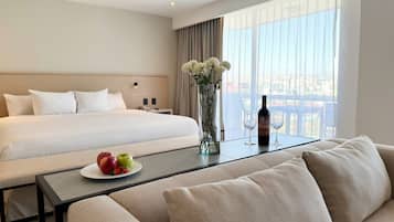 Habitación de hotel con una cama grande, un sofá, una mesa con una botella y vasos, y un florero con flores.
