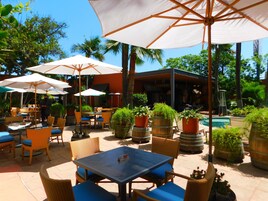 An outdoor patio area with tables and chairs under large umbrellas, surrounded by potted plants and palm trees.