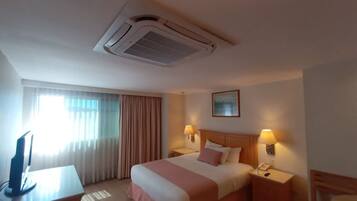 A hotel room with a bed, bedside lamps, a desk, and an air conditioning unit.