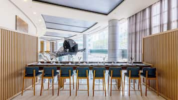 A modern dining area with a long table, multiple chairs, and a view of the city through large windows.