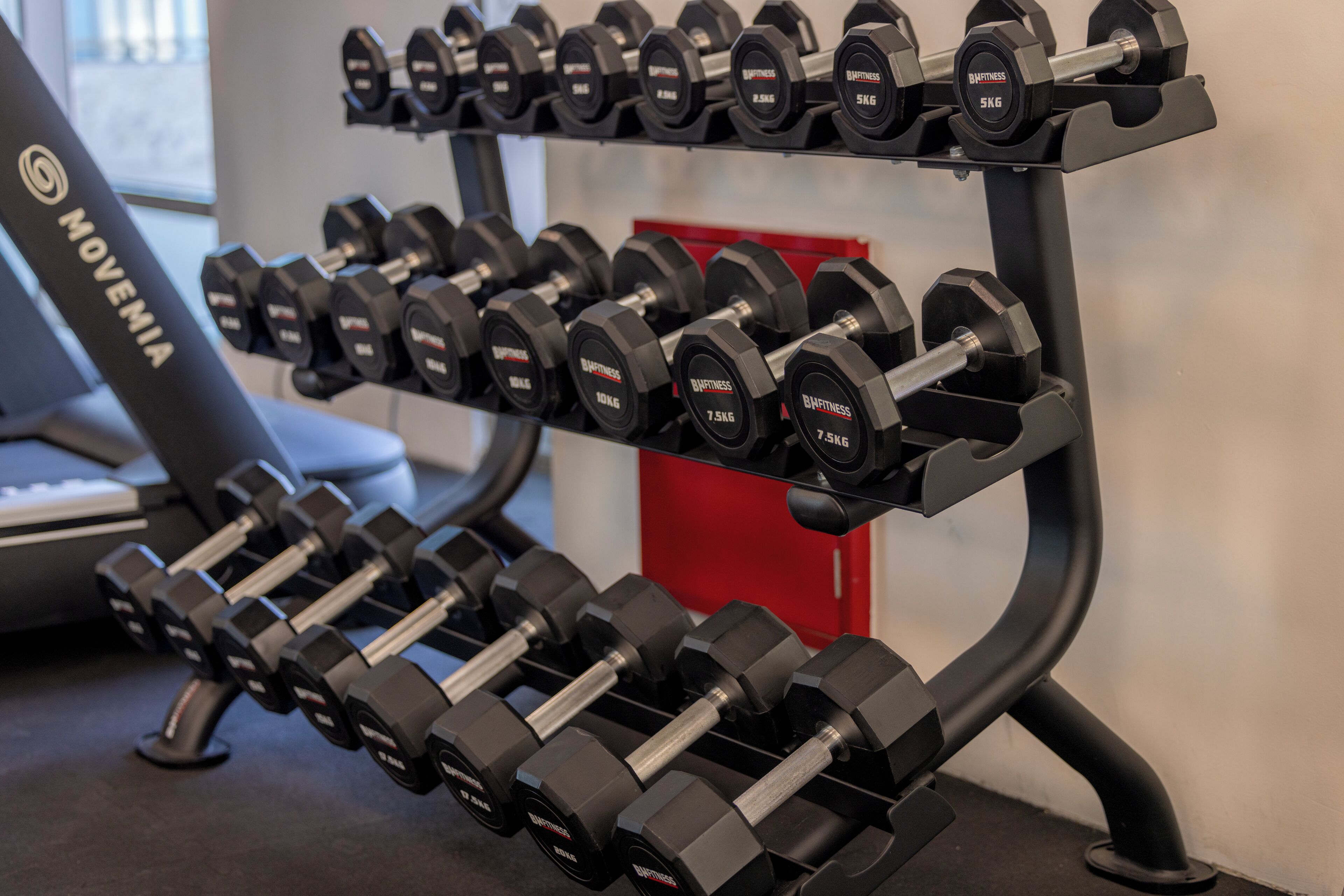 A rack of black dumbbells with weight markings, a treadmill, and a red storage unit in a gym setting.