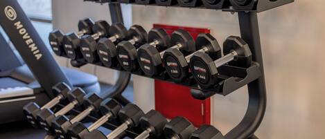 A rack of black dumbbells with weight markings, a treadmill, and a red storage unit in a gym setting.