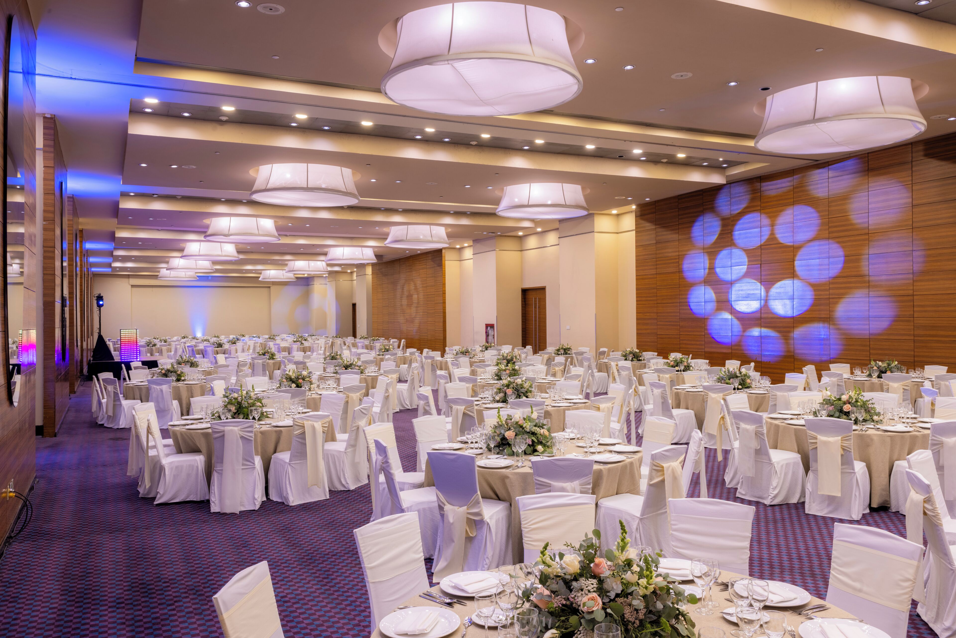 Une salle de banquet avec des tables rondes, des nappes blanches et des chaises, dotée d’un plafond avec des luminaires encastrés et d’un écran de projection affichant des motifs circulaires.