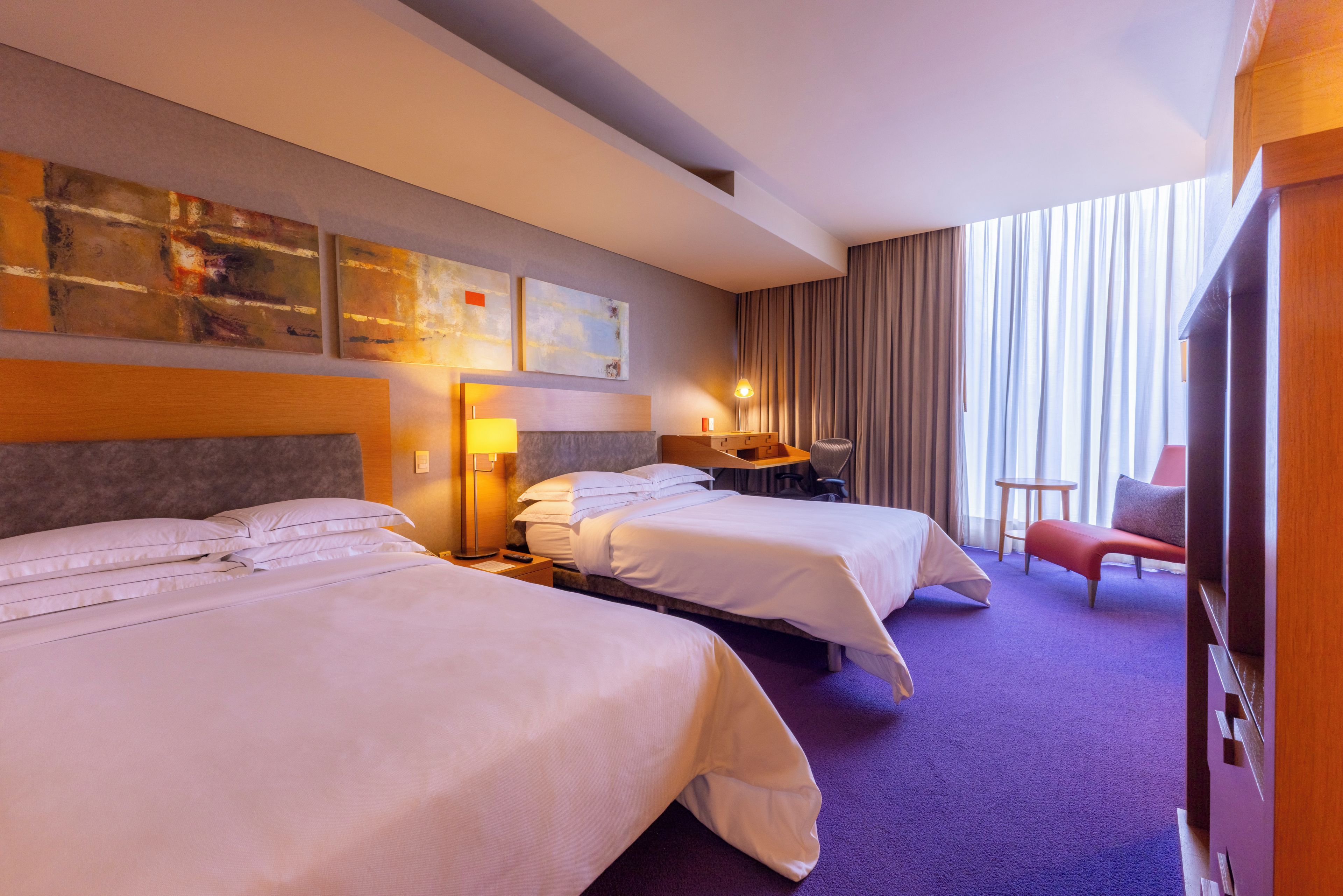 A hotel room with two beds, a desk, a chair, and a large window with curtains.
