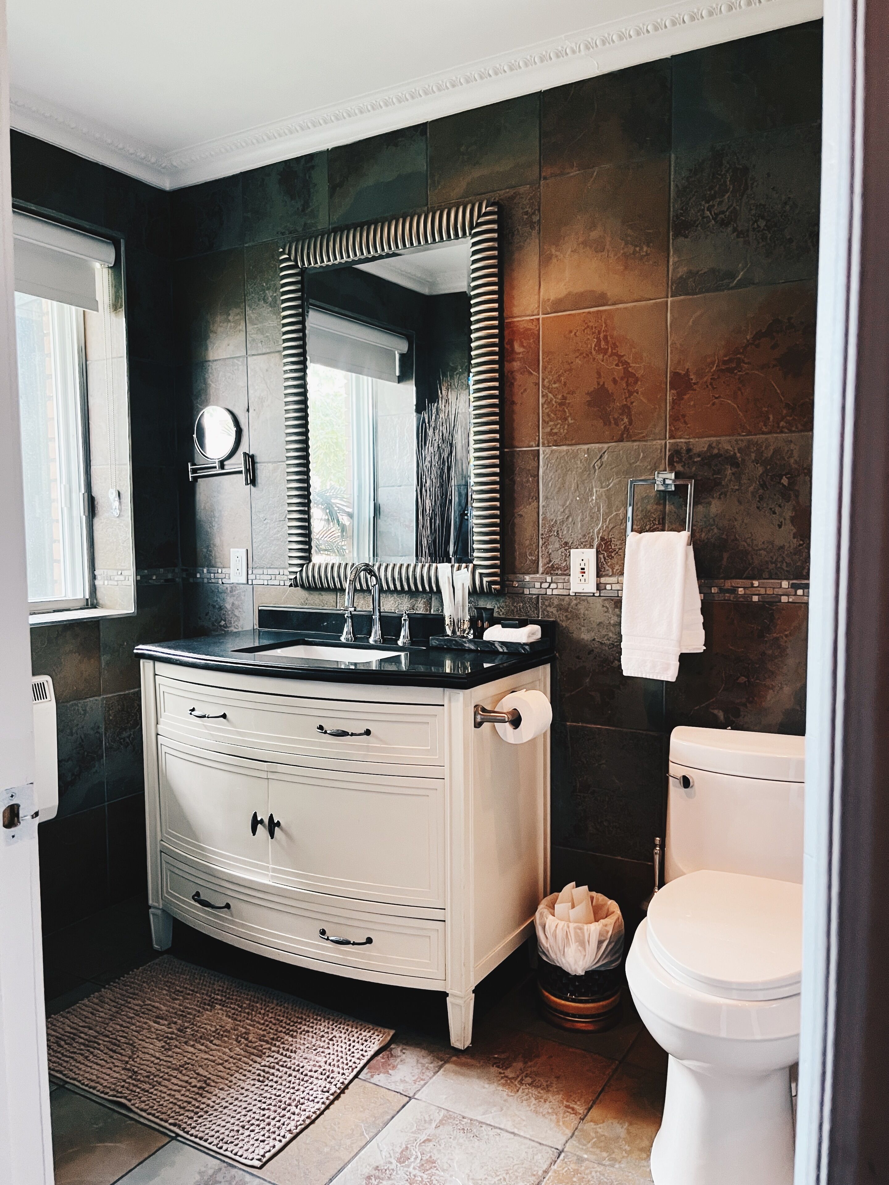 A bathroom with a large mirror, a sink, a toilet, and dark tiled walls.