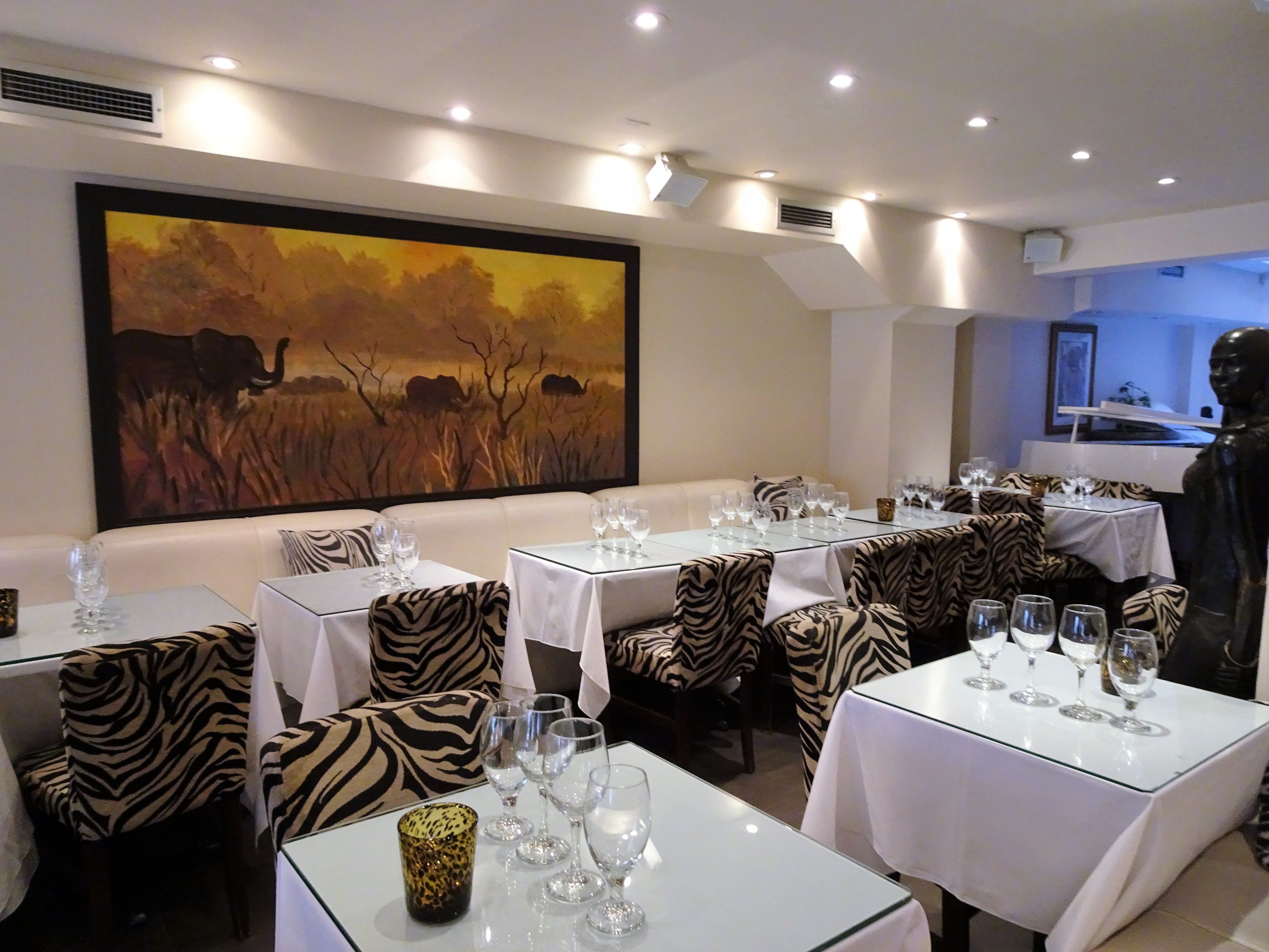 A restaurant with zebra-patterned upholstery and a large painting of an elephant in a field. Tables are set with wine glasses.