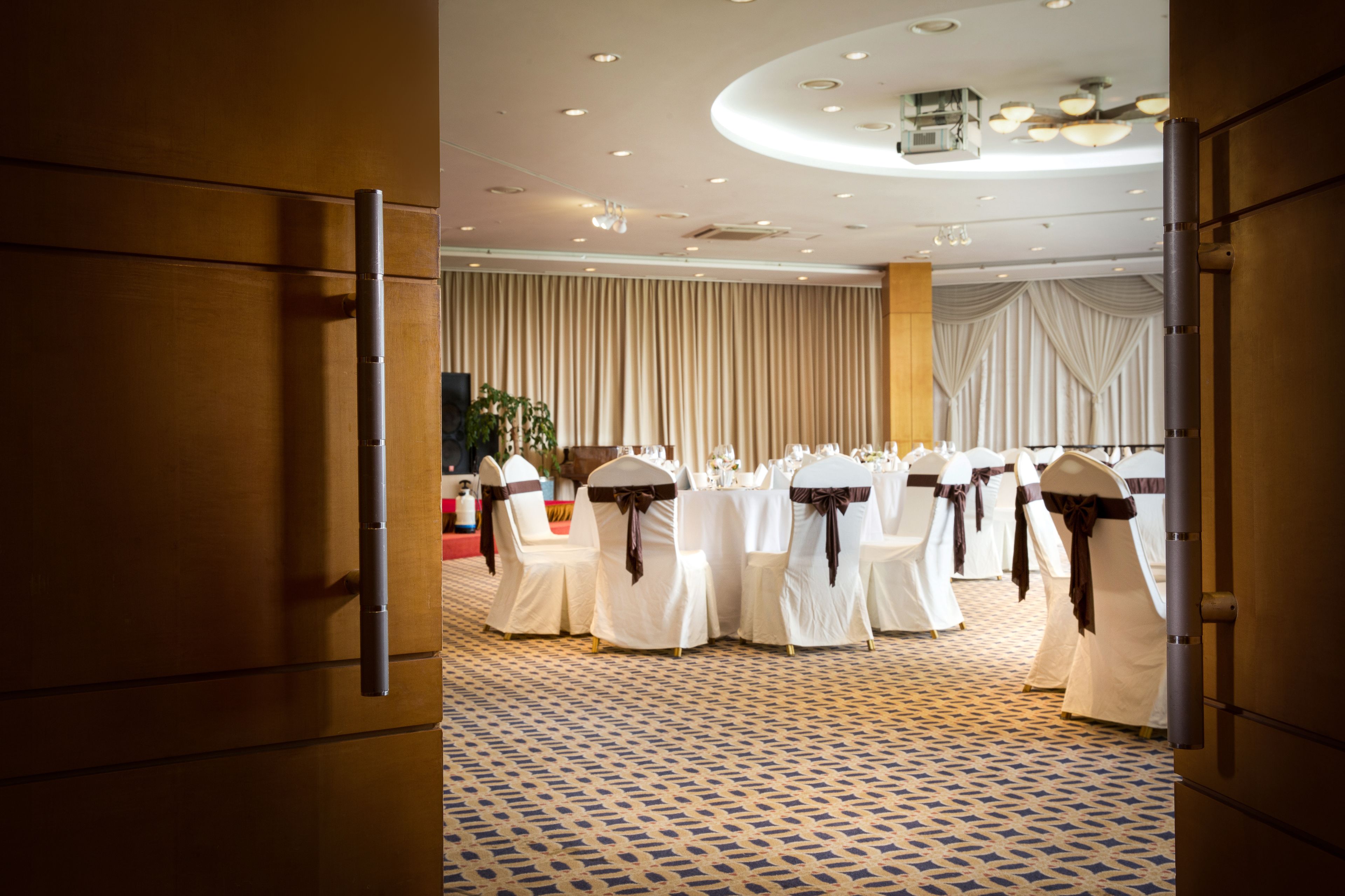 A room with a patterned carpet, white chairs with brown sashes, and a large window with curtains.
