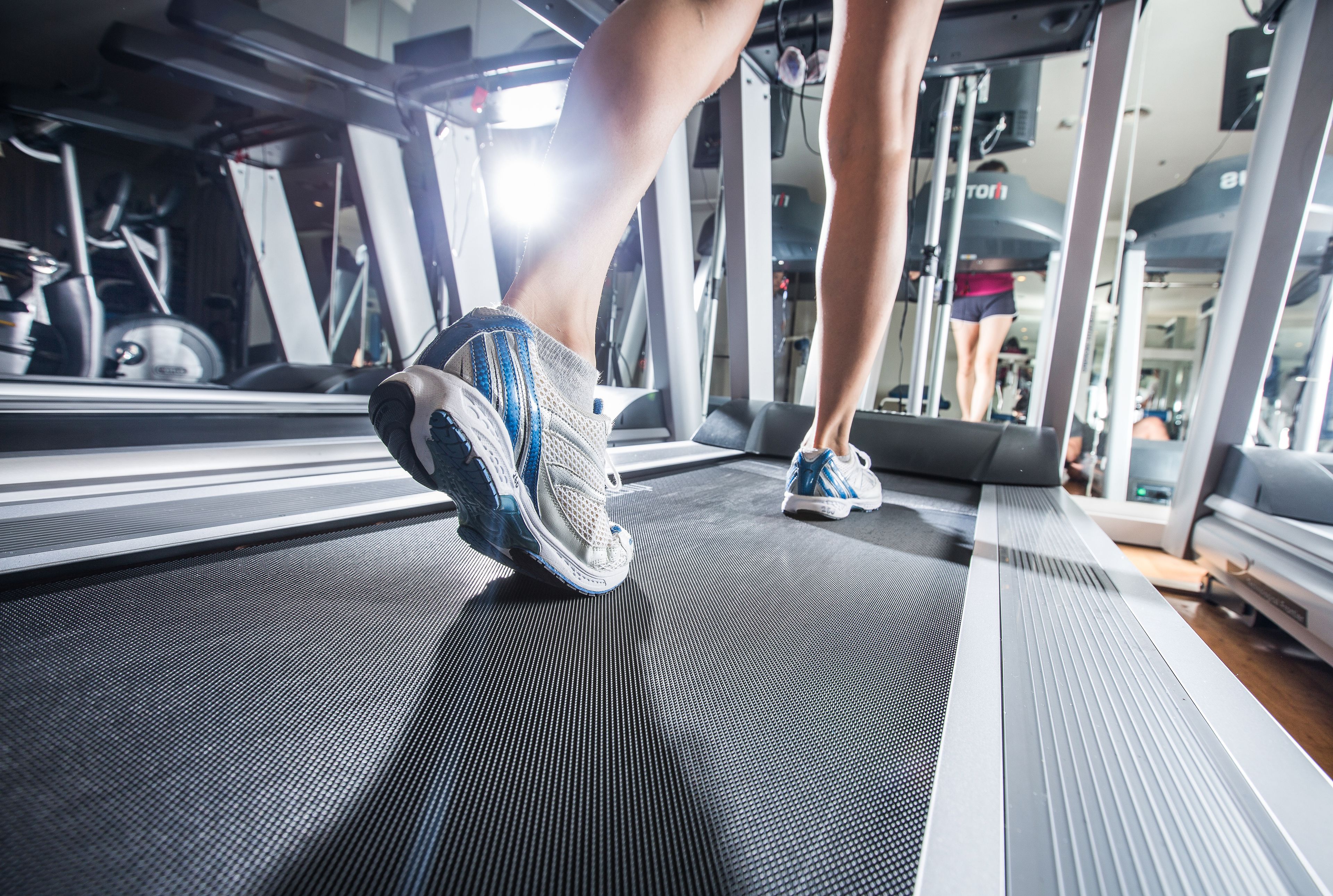 Una cinta de correr en un gimnasio con un muro espejado y equipo de ejercicio al fondo.