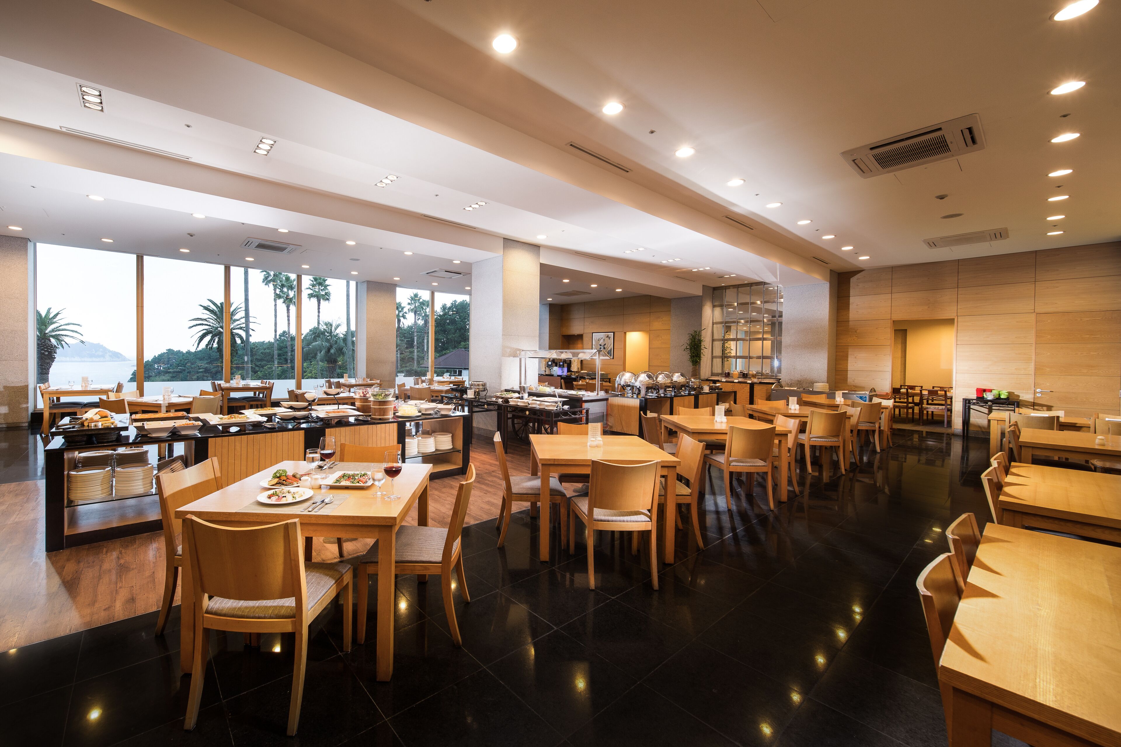 A spacious restaurant with multiple dining areas, large windows offering views of palm trees and mountains, and a modern interior design.