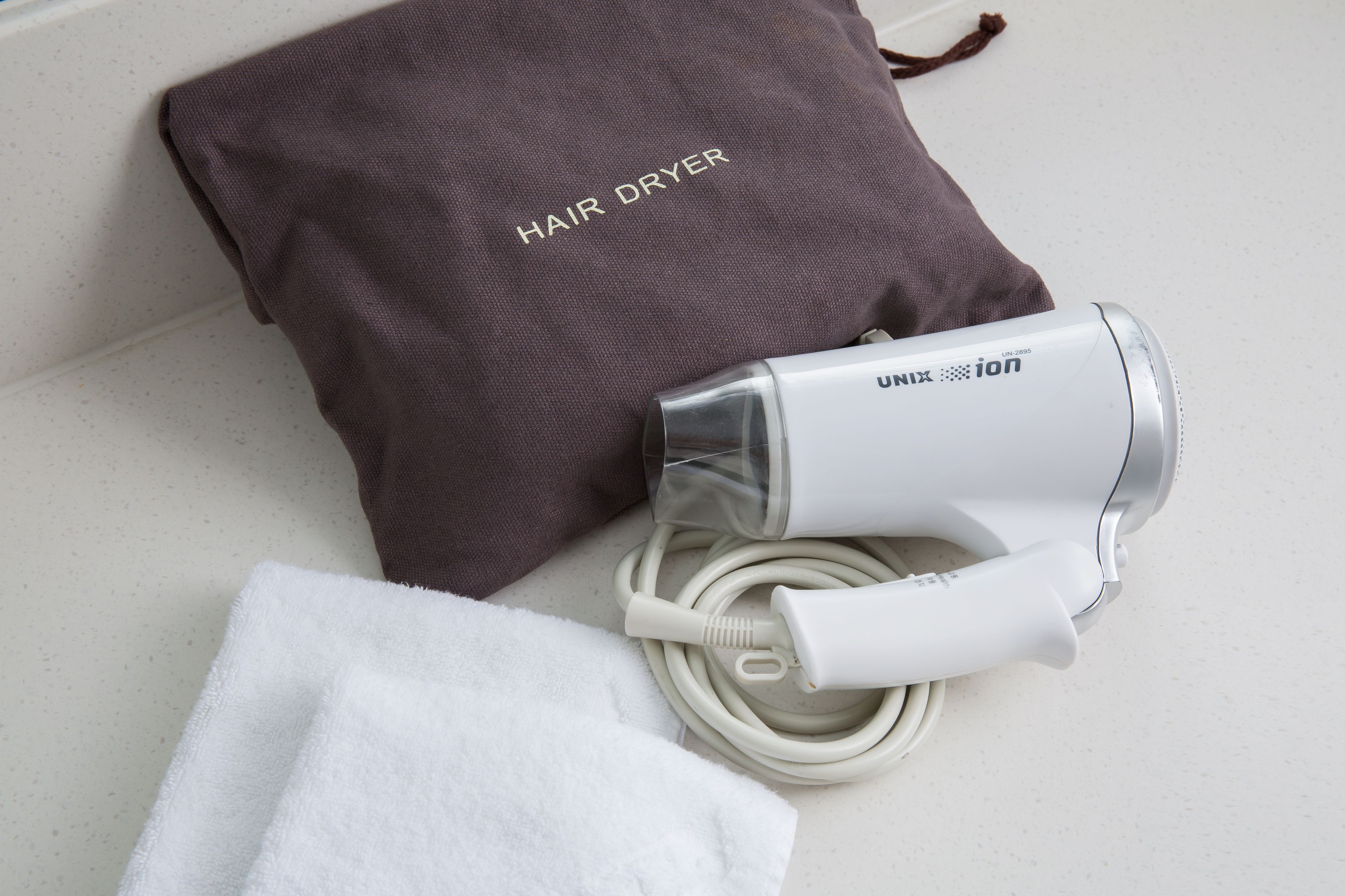 Un asciugacapelli, un asciugamano bianco e una borsa marrone con l'etichetta 'HAIR DRYER' su una superficie bianca.