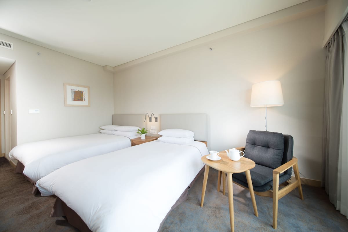 A hotel room with two single beds, a small table with a chair, a lamp, and a bedside table with a plant.