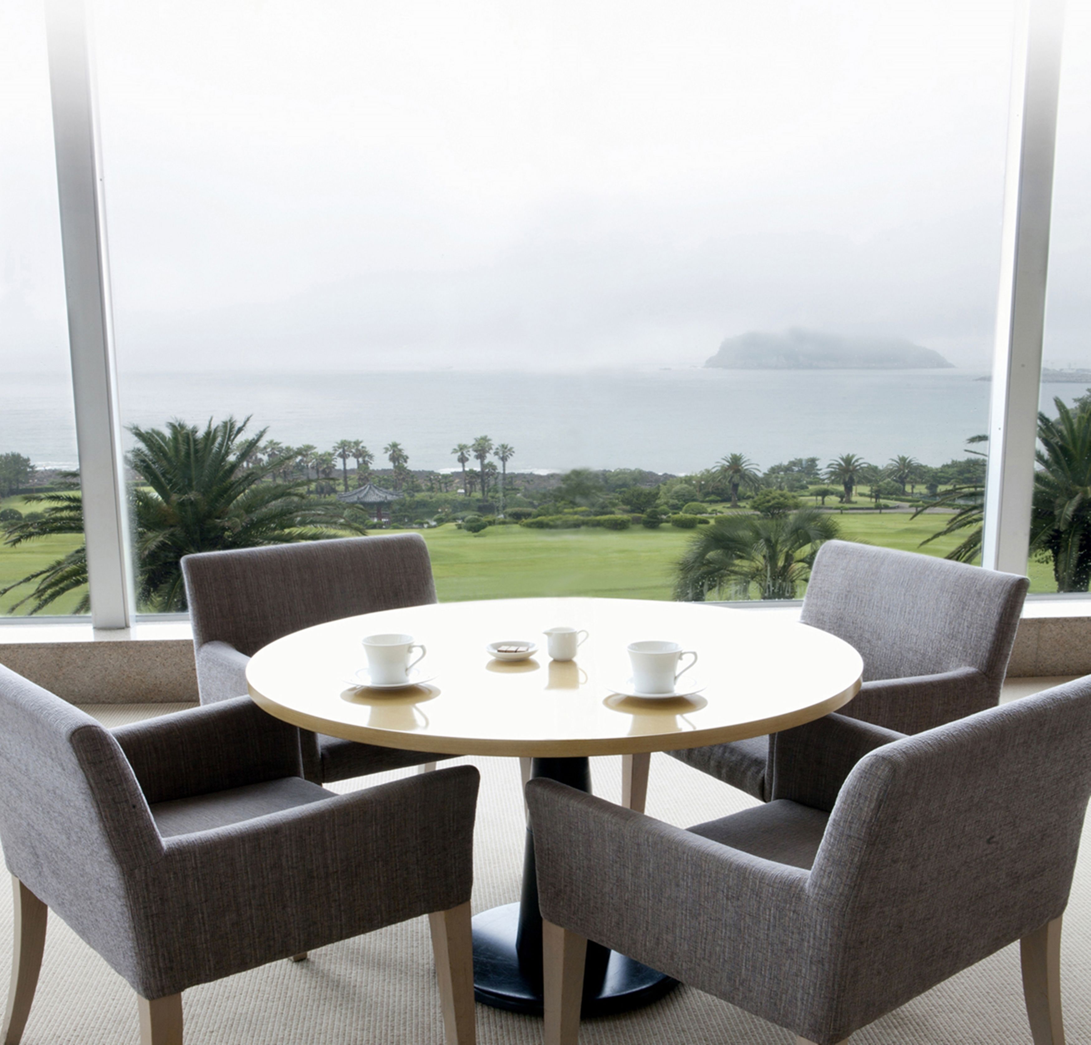 A round table with four grey chairs, a white tablecloth, and three white cups and saucers. The room has large windows offering a view of a golf course and the sea.