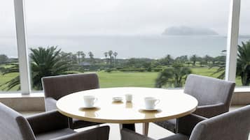 Una mesa redonda con cuatro sillas grises, un mantel blanco y tres tazas y platillos blancos. La habitación tiene ventanas grandes con vistas a un campo de golf y al mar.