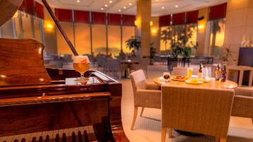 A grand piano with a glass of beer on it, a dining table with food and drinks, and a cozy seating area in a well-lit lobby.