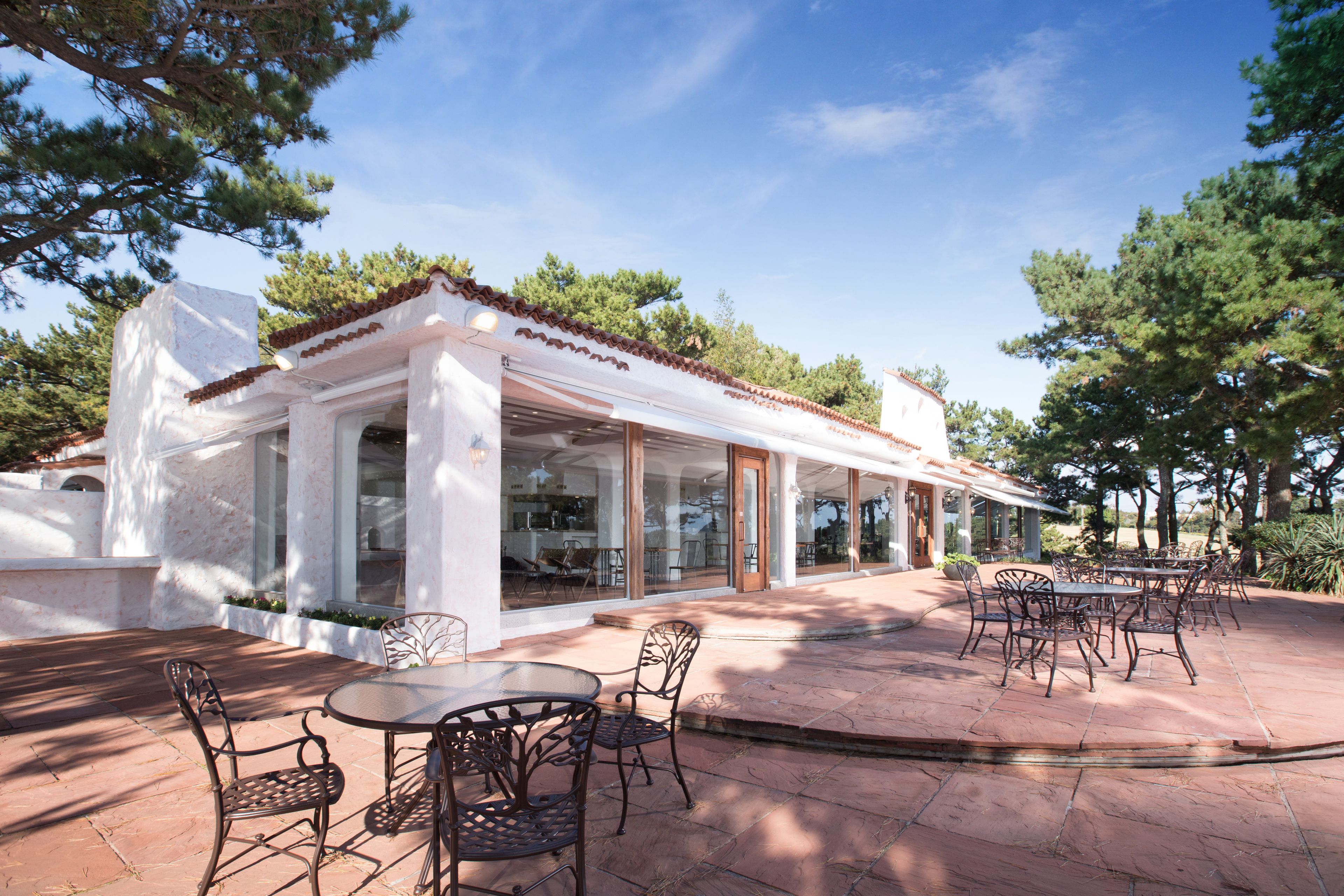 Eine überdachte Terrasse im Freien mit Tisch und Stühlen, umgeben von einem weißen Gebäude mit großen Fenstern und einem Ziegeldach.