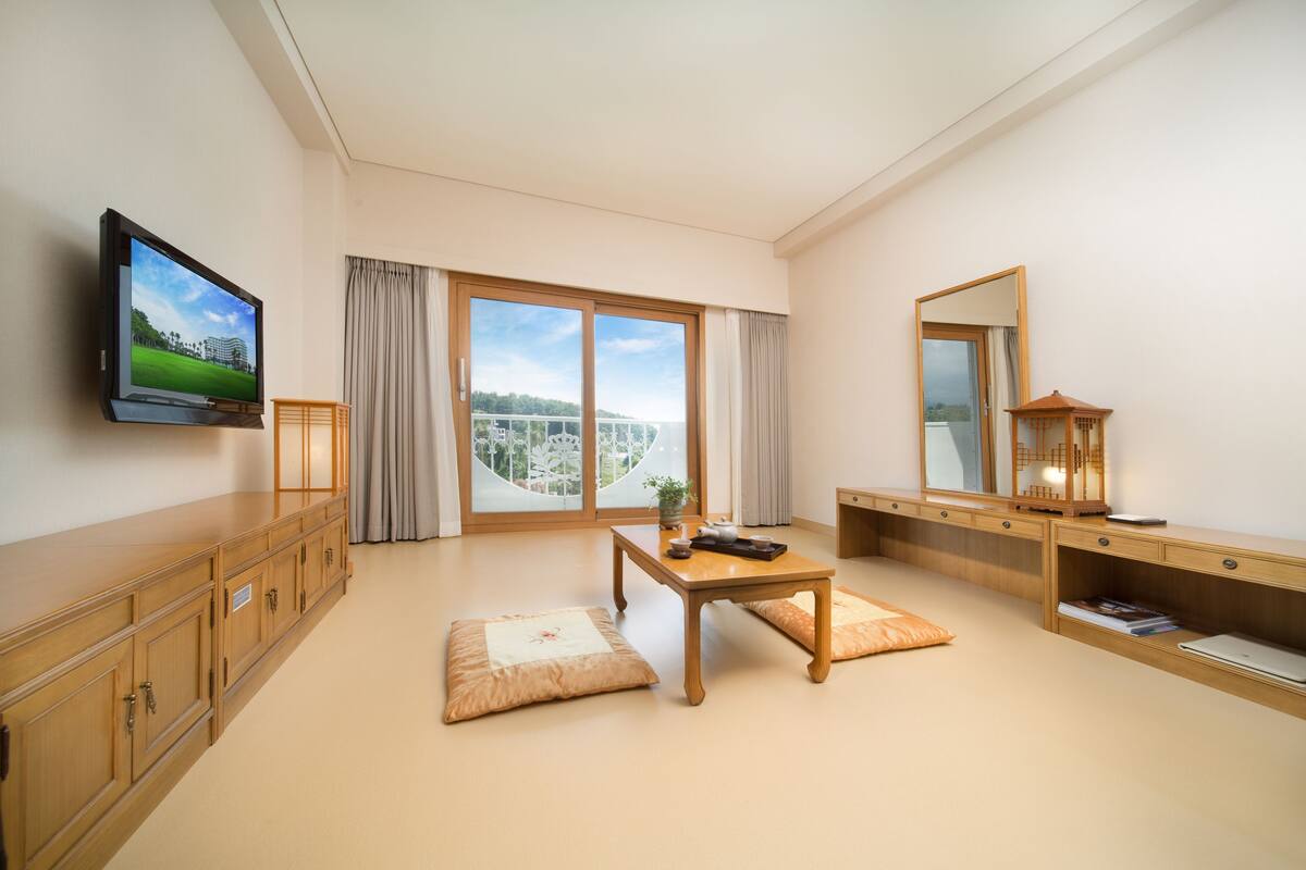 A modern hotel room with a large window, a flat-screen TV, a wooden desk, and a traditional Japanese-style lamp.