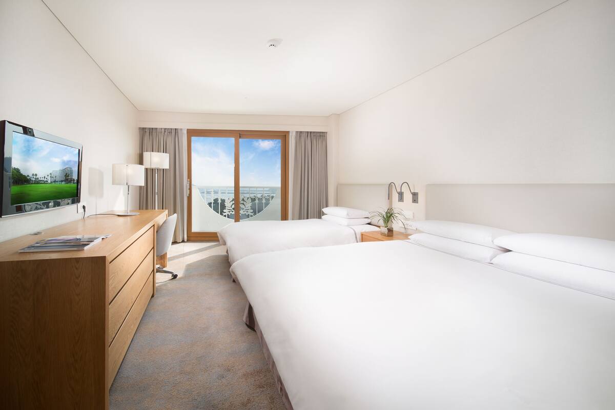A hotel room with a wooden desk, a flat-screen TV, two beds, a large window with a view, and a small plant on the nightstand.