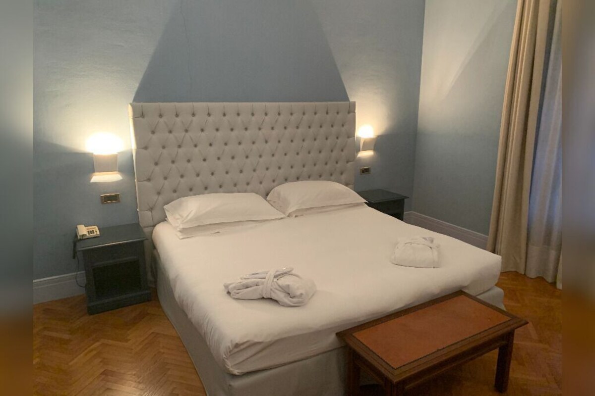 Una habitación de hotel con un cabecero capitonado, una cama individual con ropa blanca, una mesita de noche con lámpara y un banco de madera.
