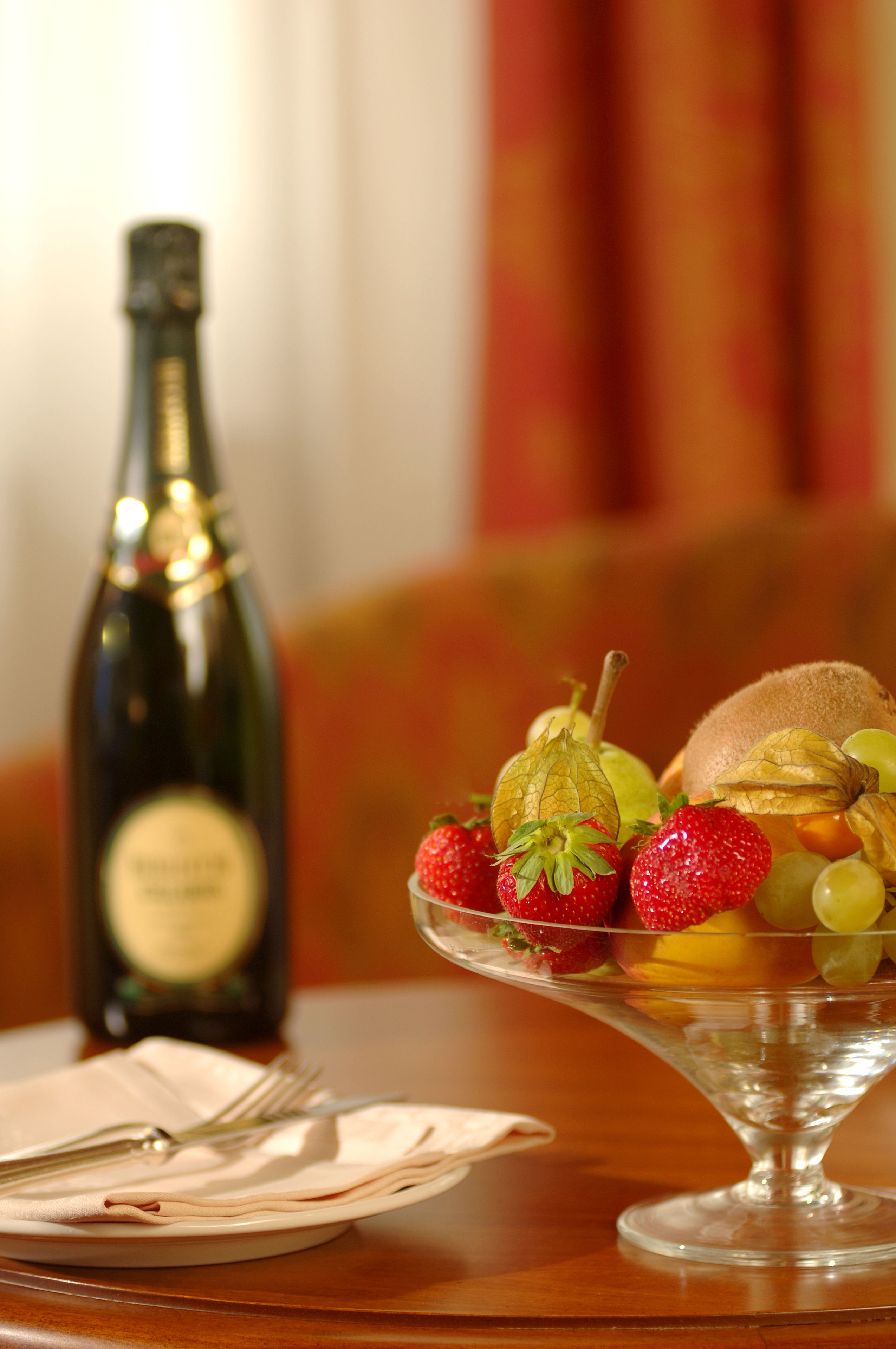Une table à manger avec un saladier en verre contenant des fraises, des raisins et des kiwis, une bouteille de vin et une serviette pliée sur laquelle repose une fourchette.