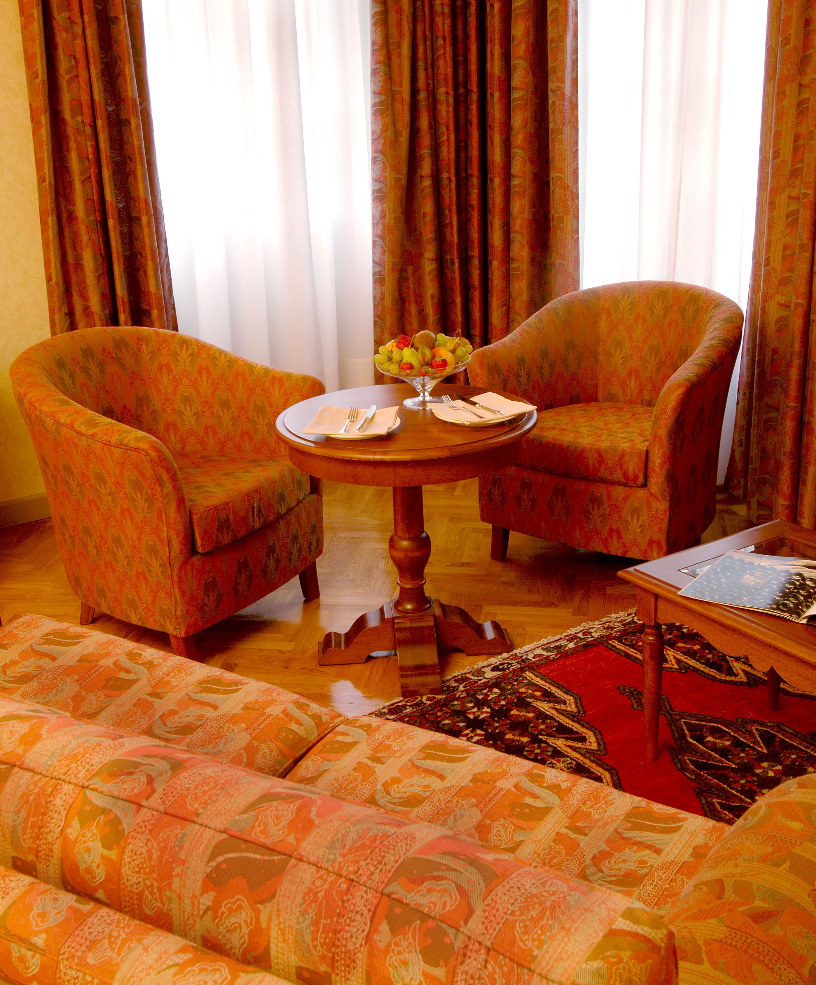 Un salon avec deux fauteuils à motifs, une table basse ronde en bois sur laquelle se trouve un panier de fruits, et un tapis rouge à motifs.