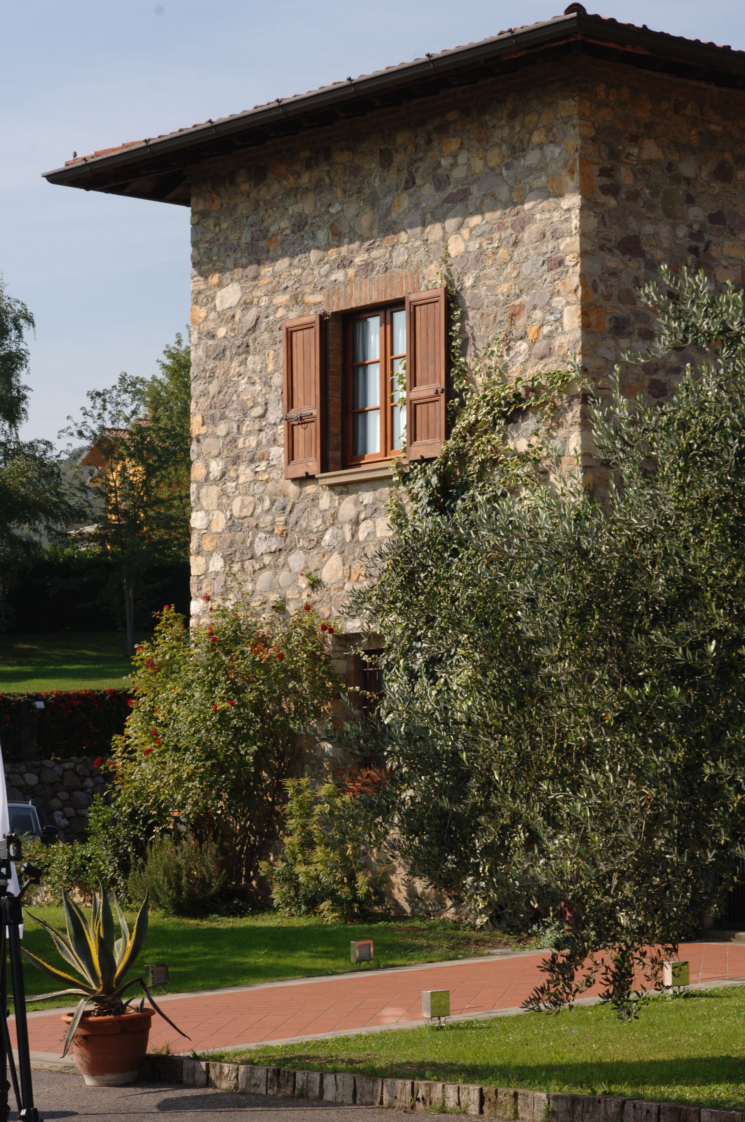 A stone building with a window and shutters, surrounded by greenery and a paved path.