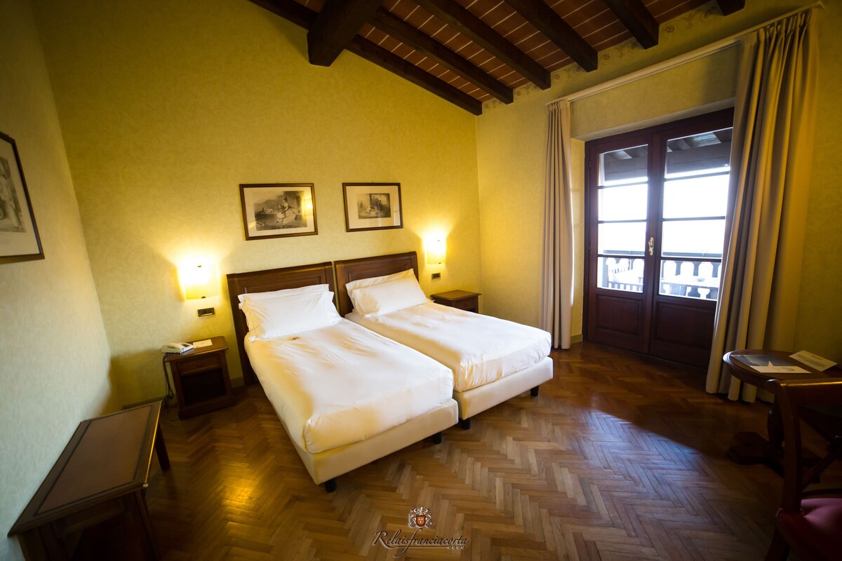 A hotel room with two beds, wooden flooring, a balcony with curtains, and framed pictures on the wall.