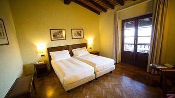 Habitación de hotel con dos camas, piso de madera, balcón con cortinas e imágenes enmarcadas en la pared.