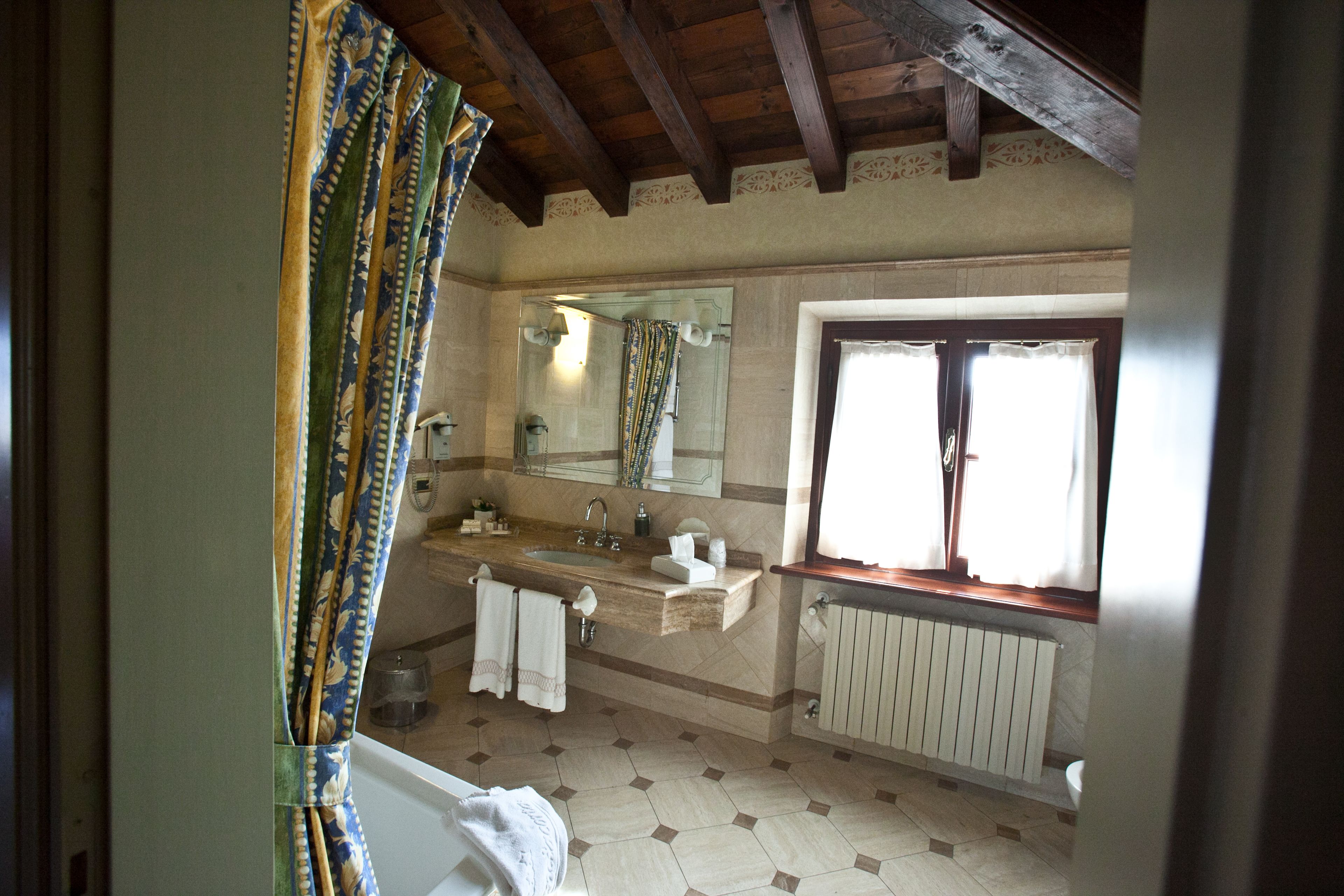 A bathroom with a wooden ceiling, a large window with curtains, a sink, a mirror, and a radiator.