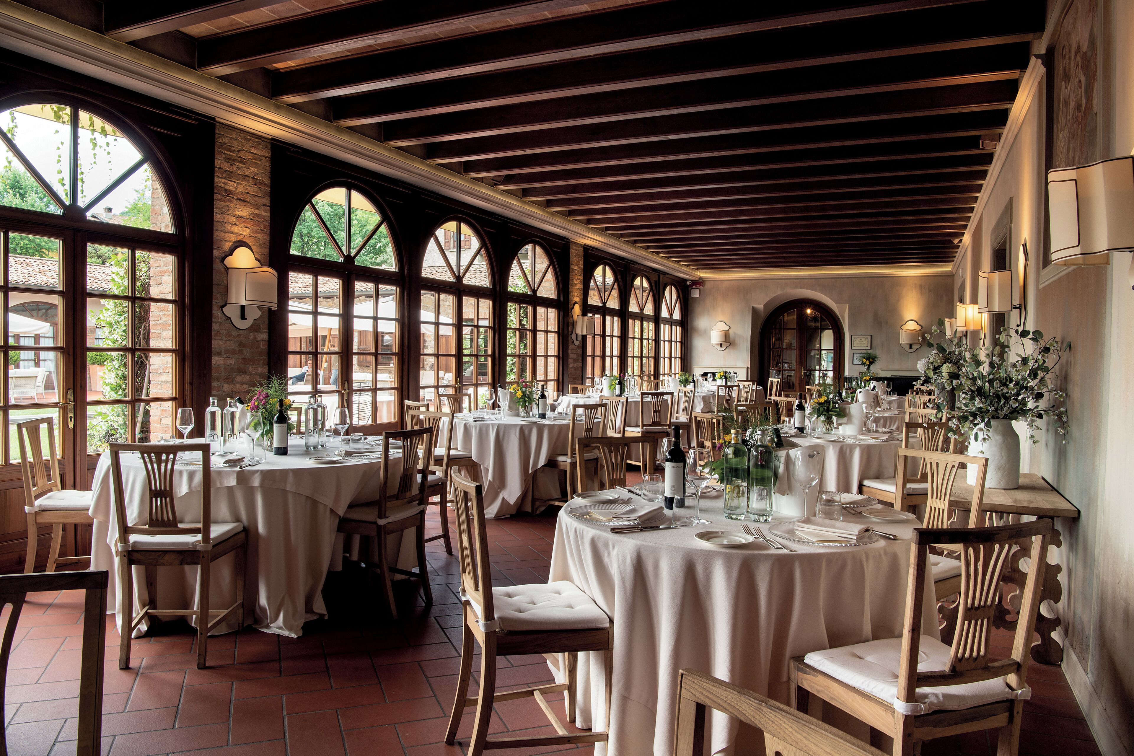 Un restaurant doté de plusieurs tables rondes, de chaises en bois et de grandes fenêtres en arc de cercle.