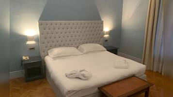 Una cama bien tendida con cabecera capitonada, una mesita de noche con lámpara y un banco de madera.