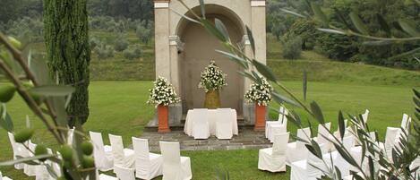 Montaje de ceremonia nupcial al aire libre con un arco de piedra, sillas blancas y arreglos florales.