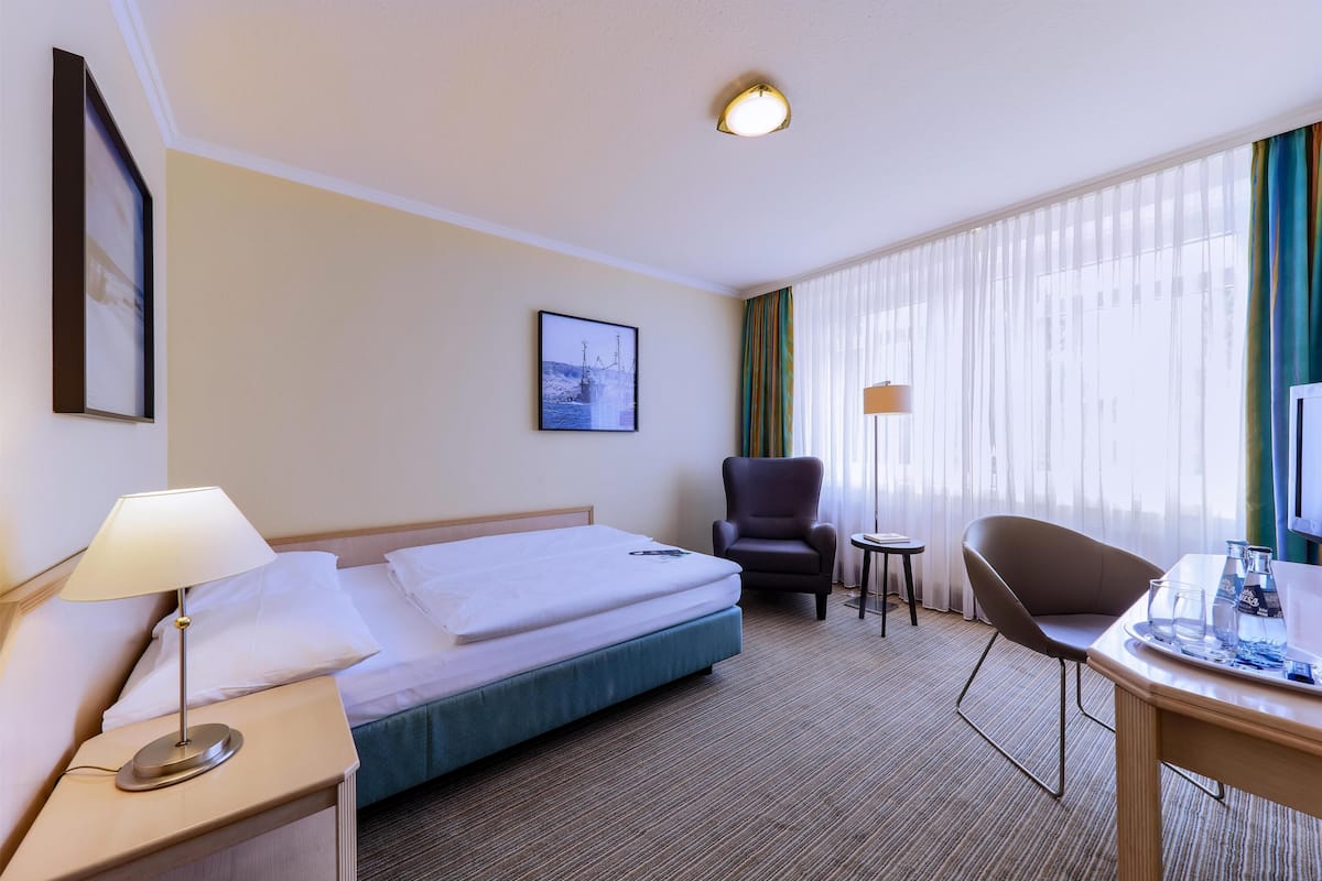A hotel room with a bed, a desk with a chair, a small table, a lamp, and a picture on the wall.
