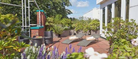A rooftop patio with two lounge chairs, a brick wall, and surrounding greenery.
