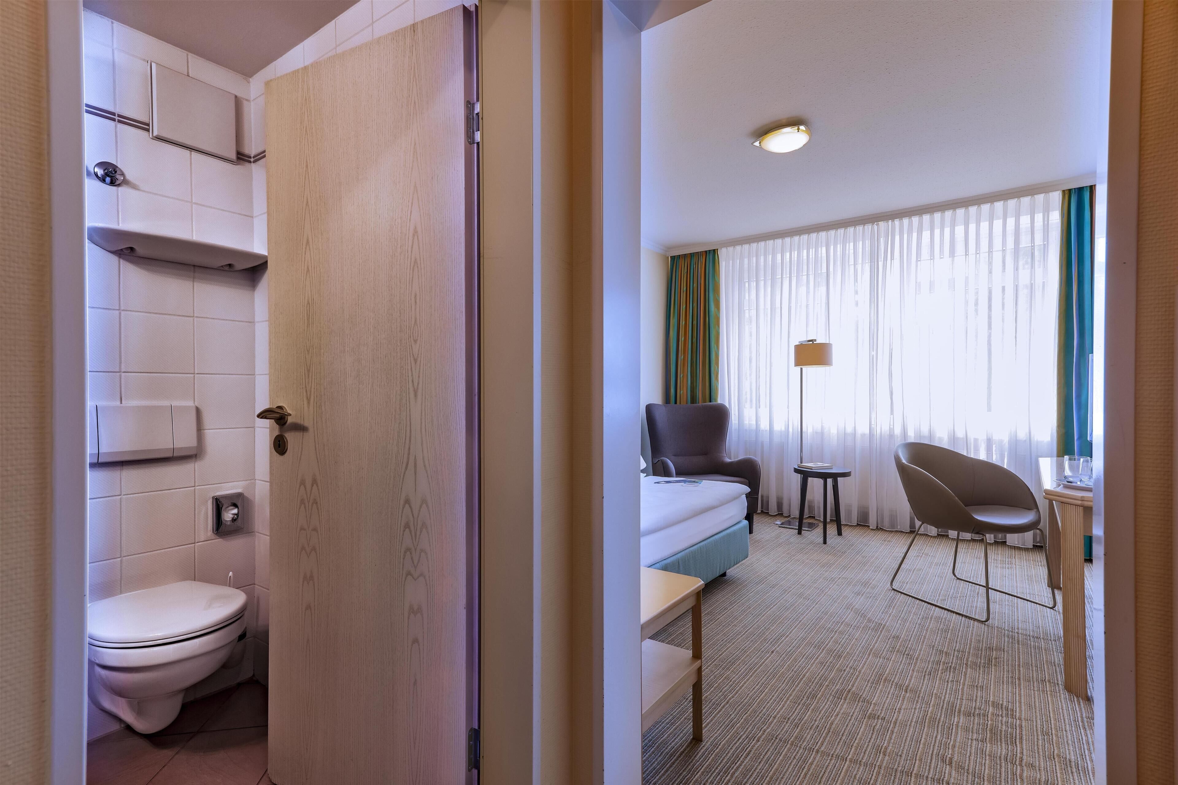 Ett hotellrum med en toalett, en säng, en stol, ett litet bord och ett fönster med gardiner.