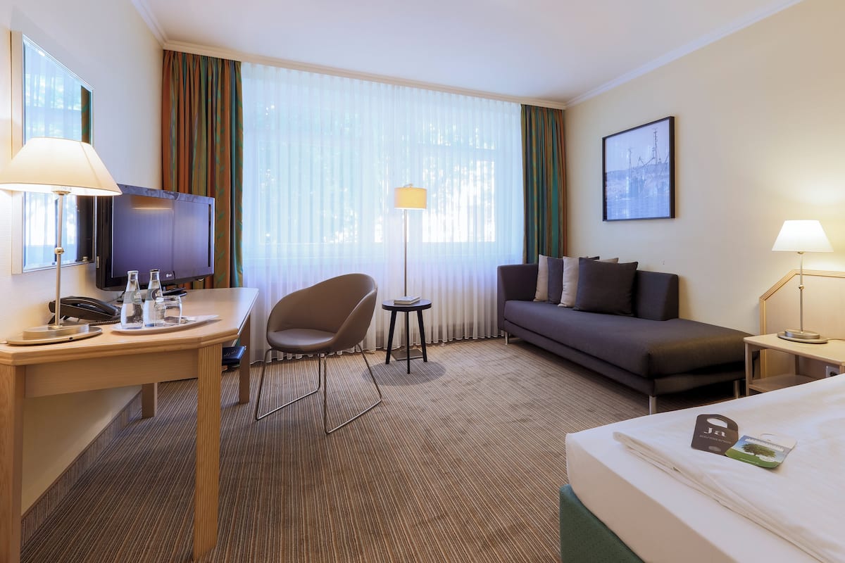 A hotel room with a bed, a desk with a lamp, a chair, a sofa, a TV, and a window with curtains.
