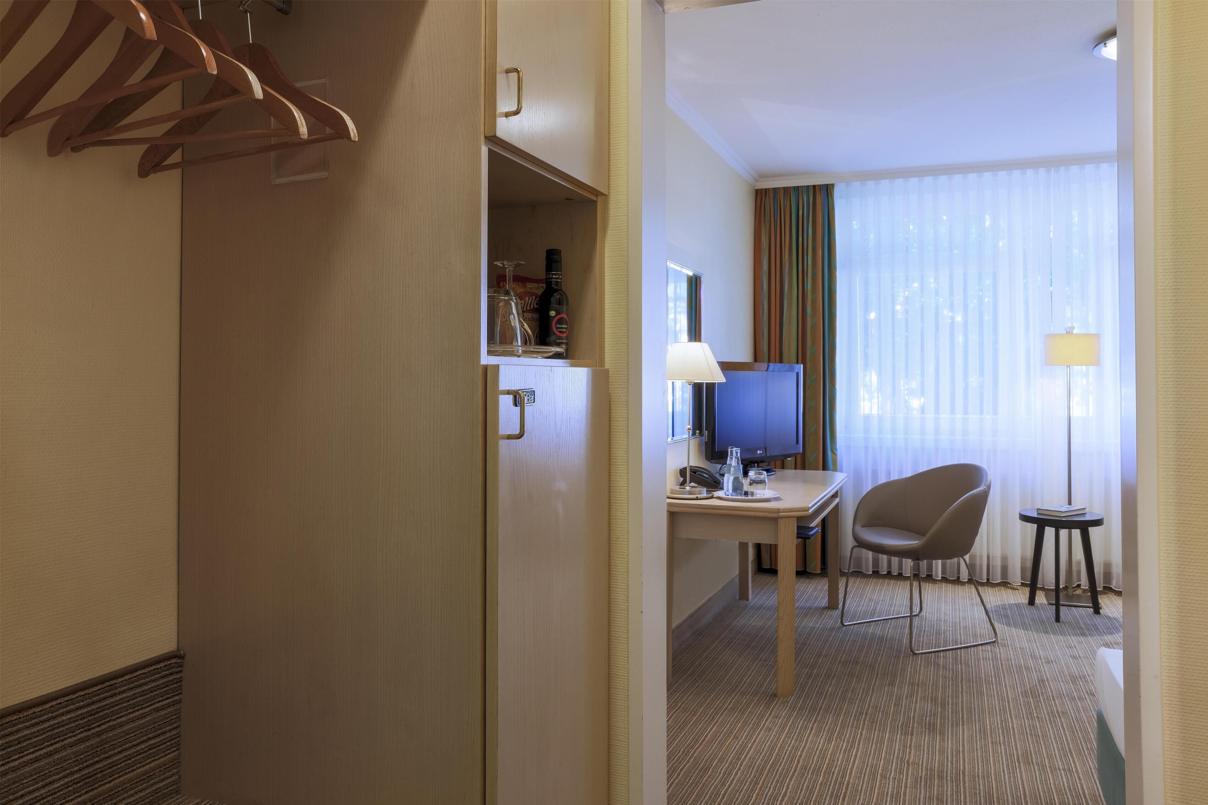 A hotel room with a desk, chair, television, and a wardrobe with hanging clothes.