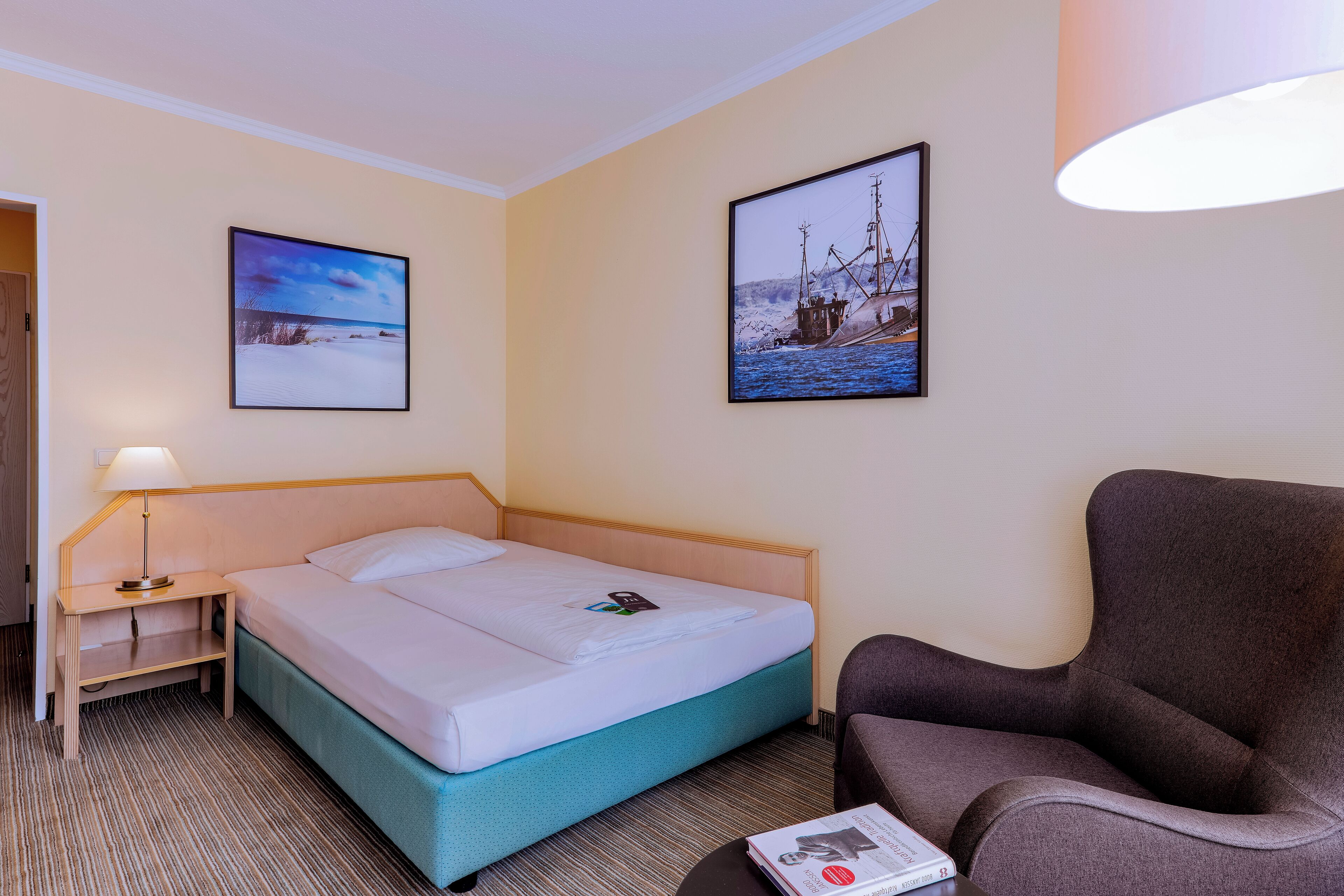 A hotel room with a bed, a chair, a nightstand with a lamp, and two framed pictures on the wall.