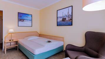 A hotel room with a bed, a chair, a nightstand with a lamp, and two framed pictures on the wall.