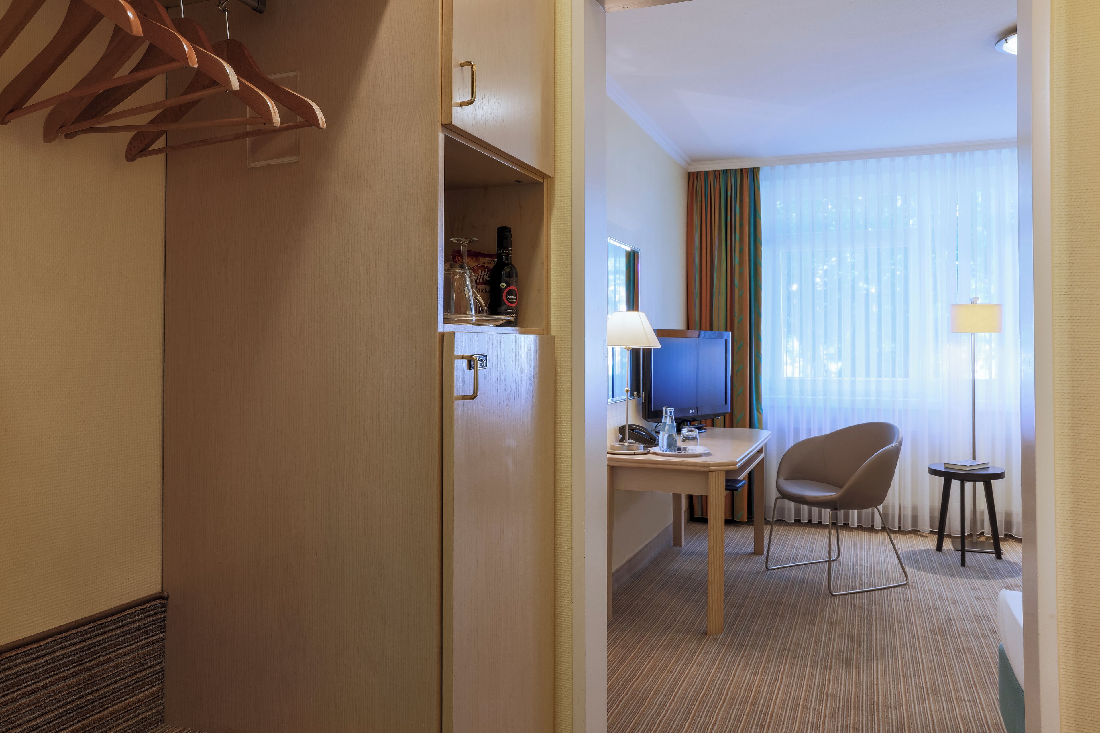 A hotel room with a wooden wardrobe, a desk with a chair, a television, and a lamp.