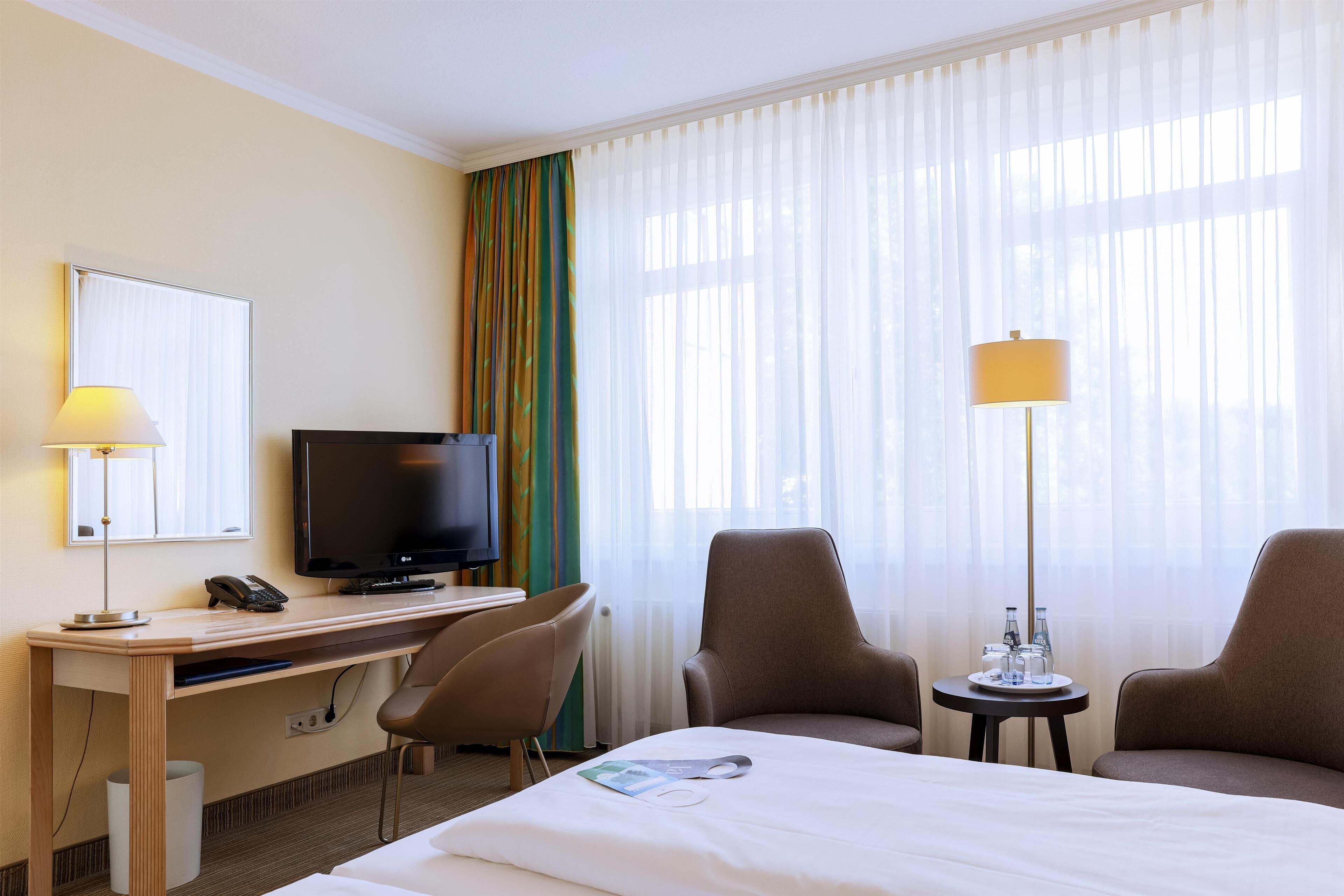 A hotel room with a bed, a desk with a TV, two chairs, a lamp, and a window with sheer curtains.