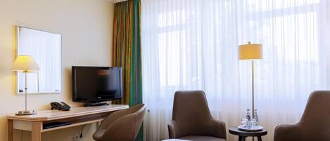 Habitación de hotel con cama, escritorio con televisor, dos sillas, lámpara y ventana con cortinas traslúcidas.