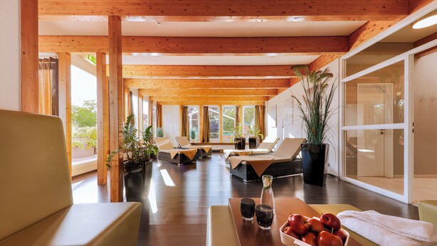 Un espacio interior amplio con vigas de madera, asientos cómodos, una mesa con frutas y bebidas, y ventanas grandes.
