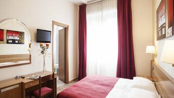 Quarto de hotel com cama, secretária e cadeira, espelho, televisão e janela com cortinas.