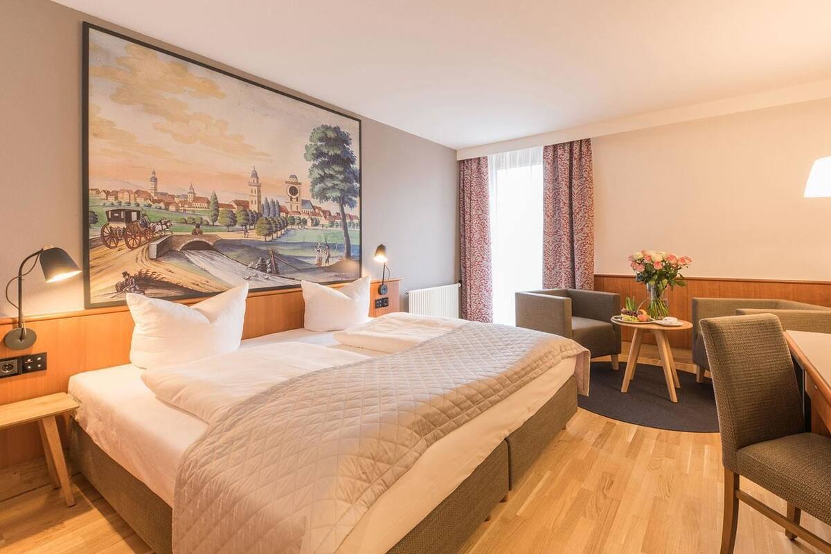 A hotel room with a large bed, a desk, a chair, a table with flowers, and a painting on the wall.