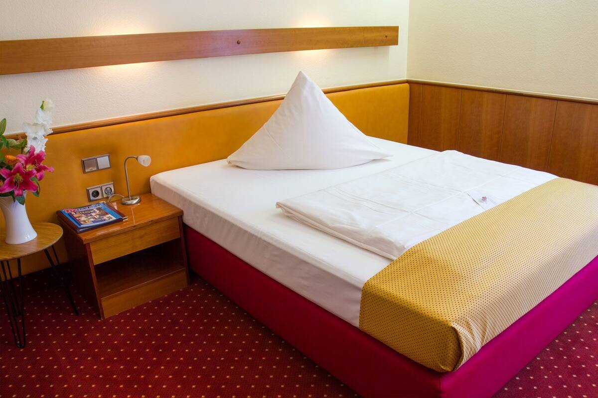 A hotel room with a wooden headboard, a single bed with white bedding, a red carpet, a small bedside table with a lamp, a magazine, and a vase with flowers.