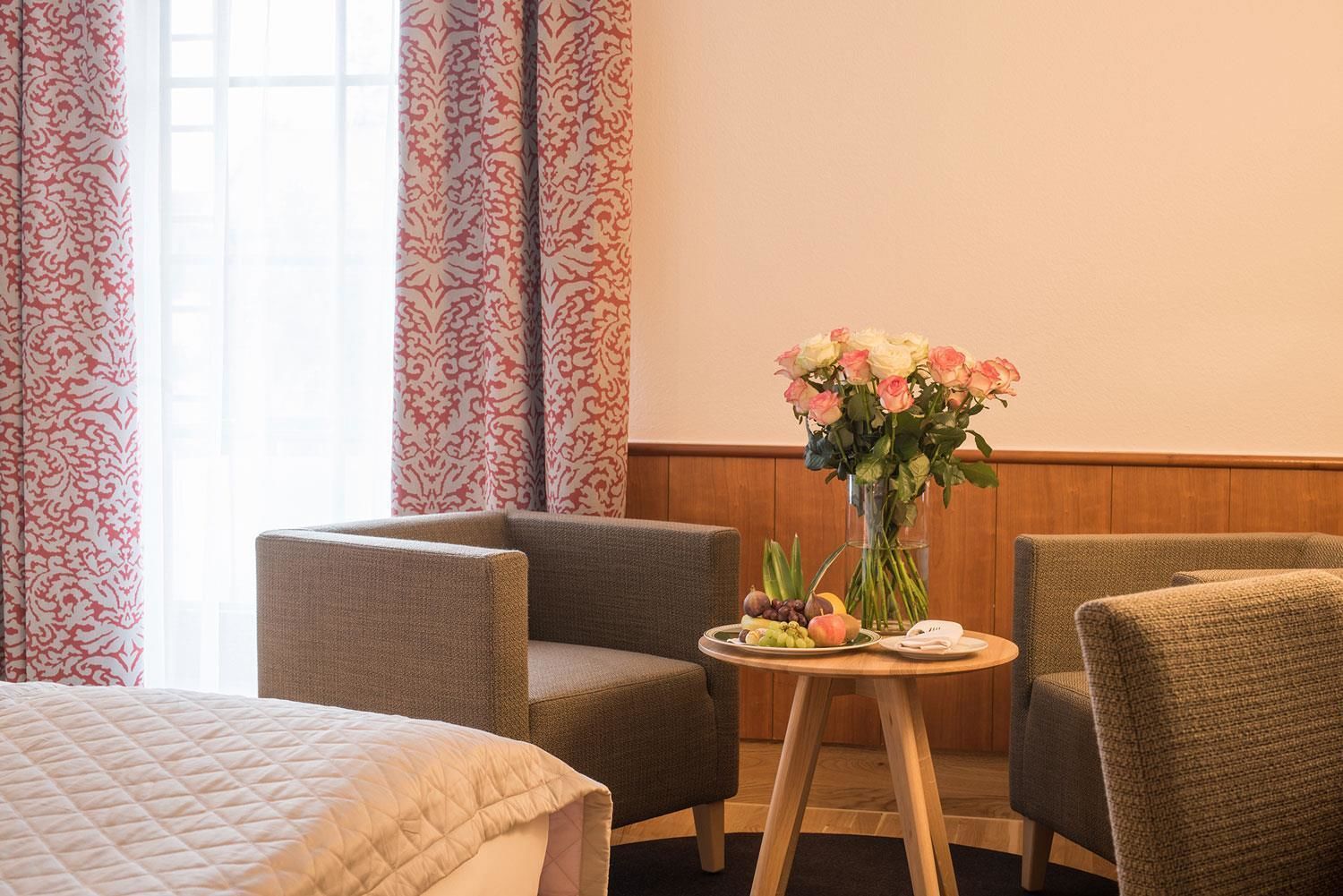 Quarto de hotel com cama, duas poltronas, uma mesinha de centro com um vaso de flores e um prato de frutas.