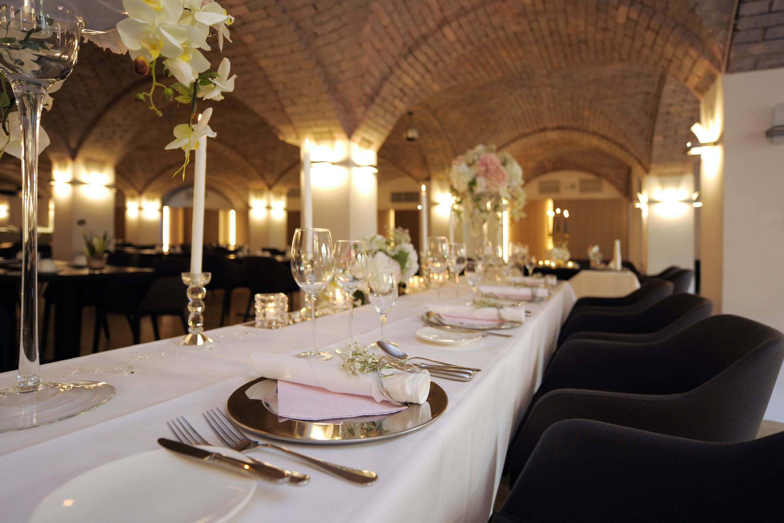 Une longue table de banquet, recouverte de nappes blanches, agrémentée d’argenterie et de verres, avec un plafond en briques et des lustres en arrière-plan.
