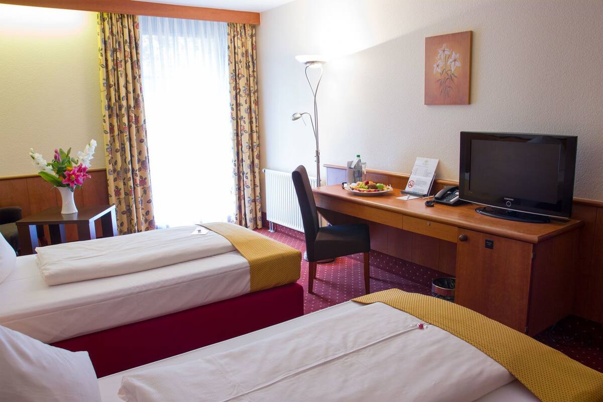 A hotel room with two beds, a desk with a computer and phone, a lamp, and a painting on the wall.
