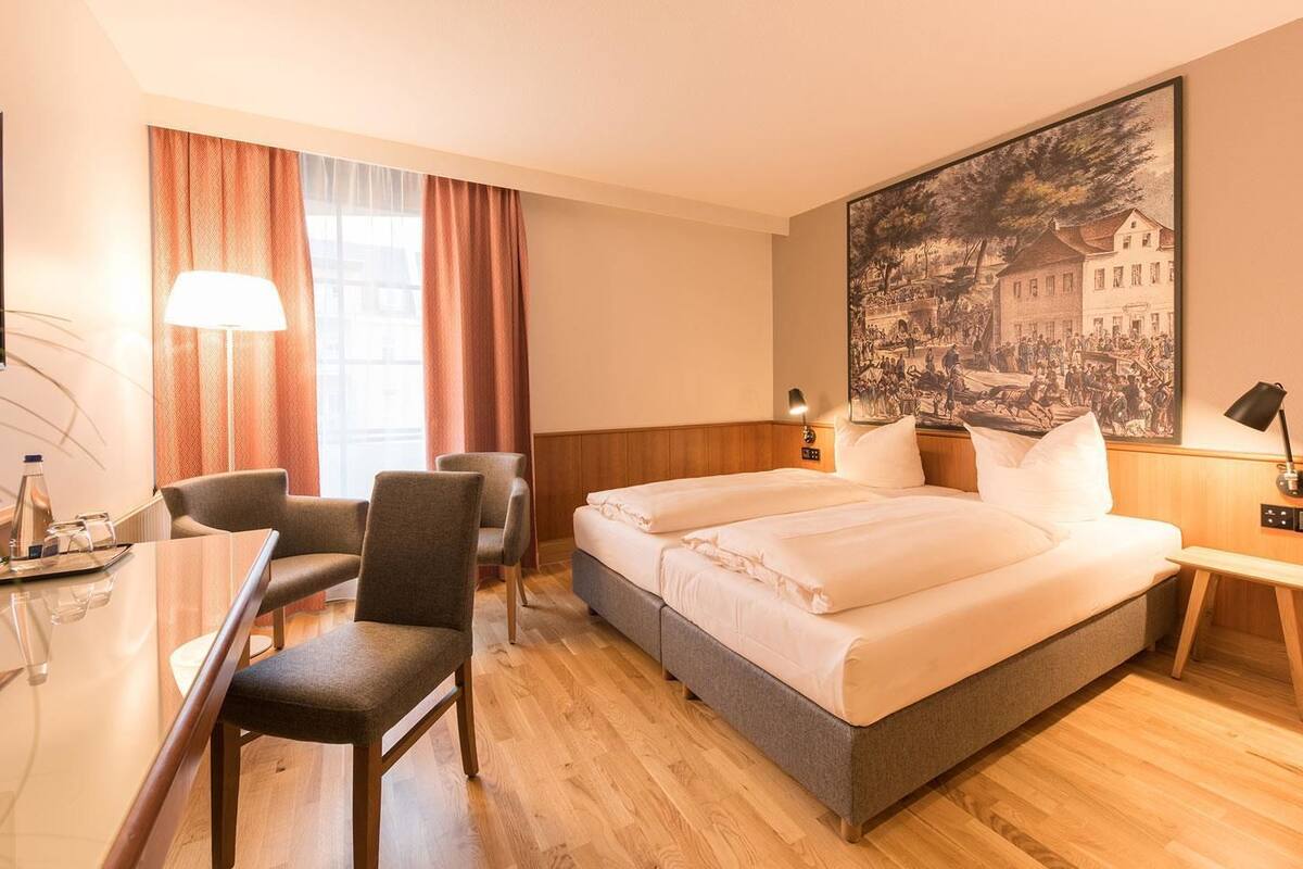 A hotel room with a bed, two chairs, a desk, a lamp, and a painting on the wall.