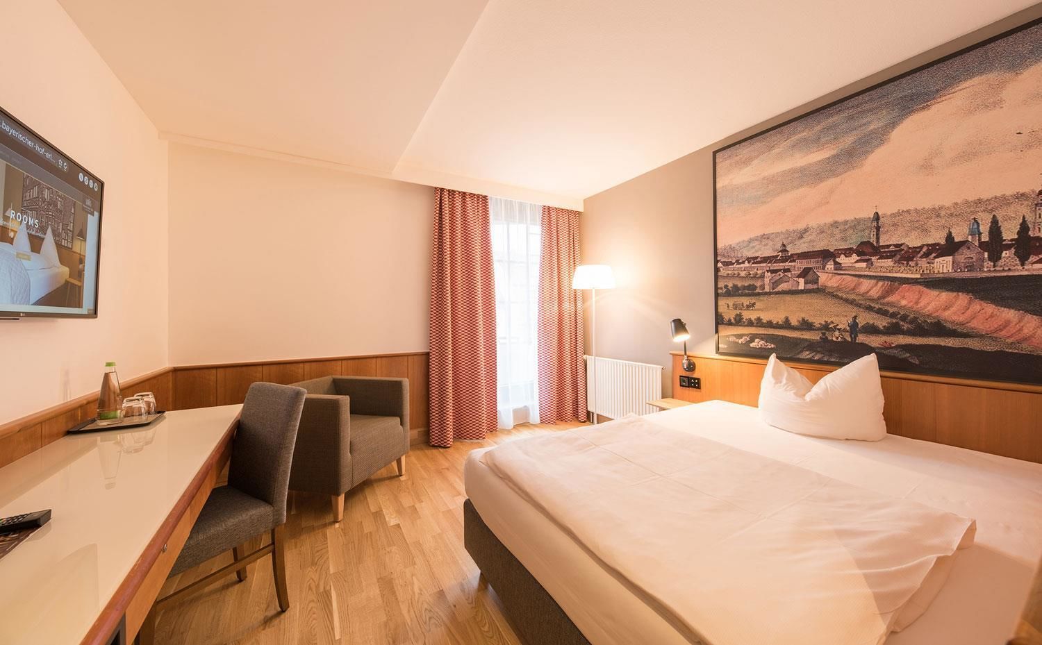 A hotel room with a bed, a desk, a chair, a lamp, and a painting on the wall.